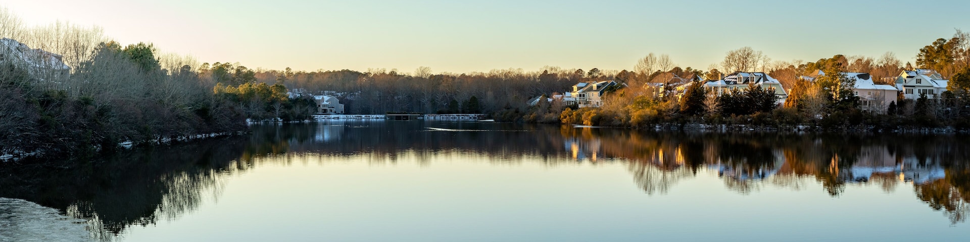A calm lake with reflections of houses and trees, surrounded by a forested area.