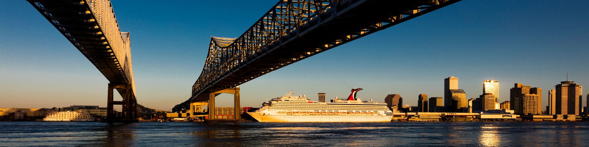 A cruise ship under a large bridge with a city skyline in the background.
