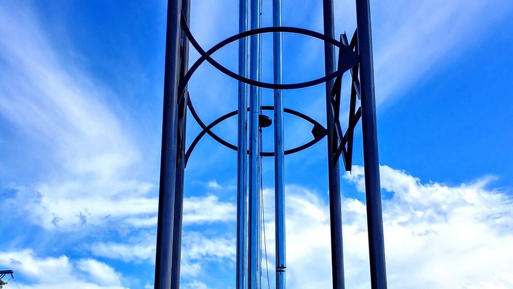 A tall, metallic sculpture with a circular base and intersecting arms, set against a clear blue sky with clouds.