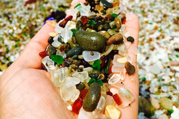 A hand holding a variety of colorful stones and crystals by a pebble beach.