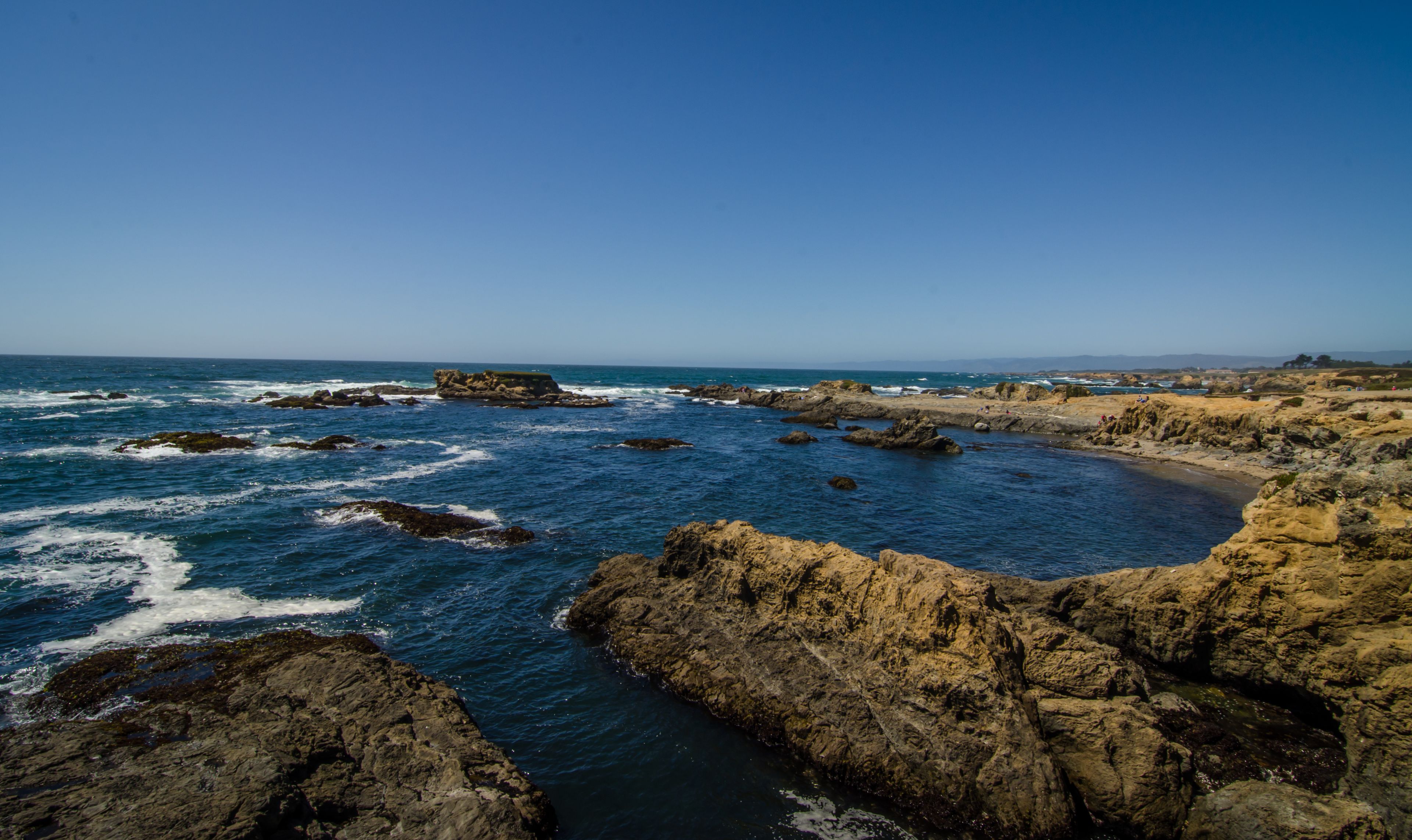 A rocky coastline with waves crashing against the rocks and a clear blue sky.