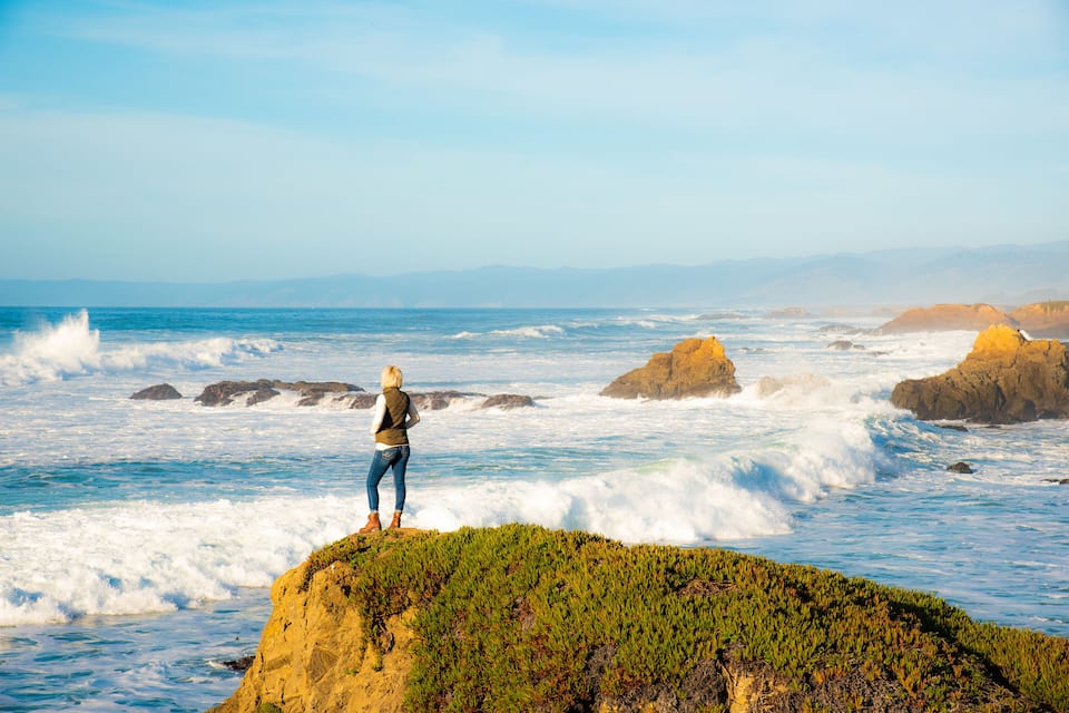 Une personne se tient debout sur une falaise, surplombant l’océan, tandis que les vagues s’écrasent contre les rochers.