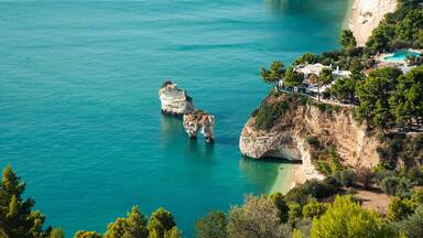 A coastal landscape with clear turquoise waters, rocky cliffs, and a small building complex on the right.