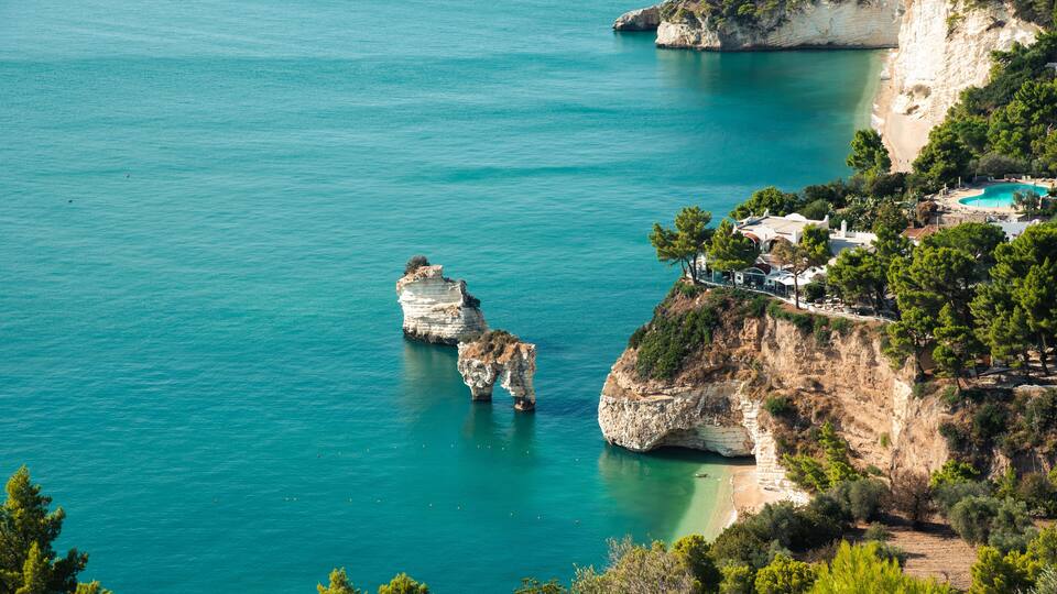 A coastal landscape with clear turquoise waters, rocky cliffs, and a small building complex on the right.