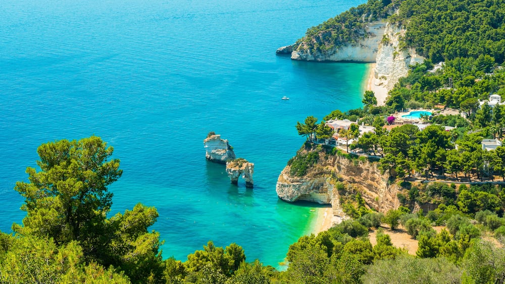 A coastal landscape with clear turquoise waters, rocky outcrops, and a beach with buildings and greenery.