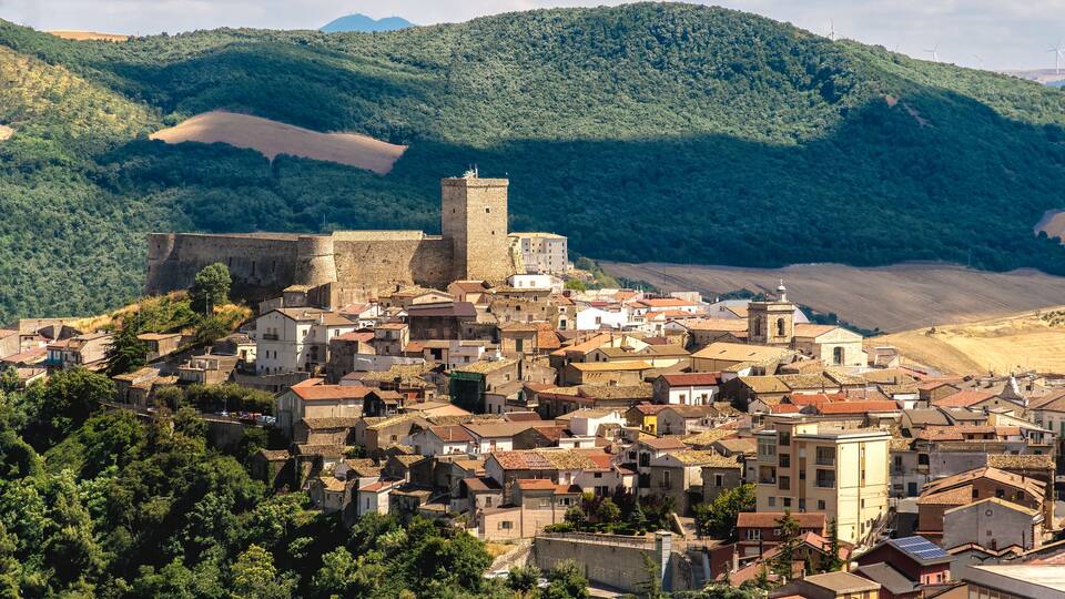 A historic town with a prominent castle, surrounded by lush green hills and wind turbines in the background.