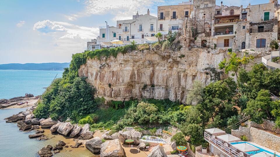 A coastal town with historic buildings perched on a cliff, a beach with large rocks, and a swimming pool.