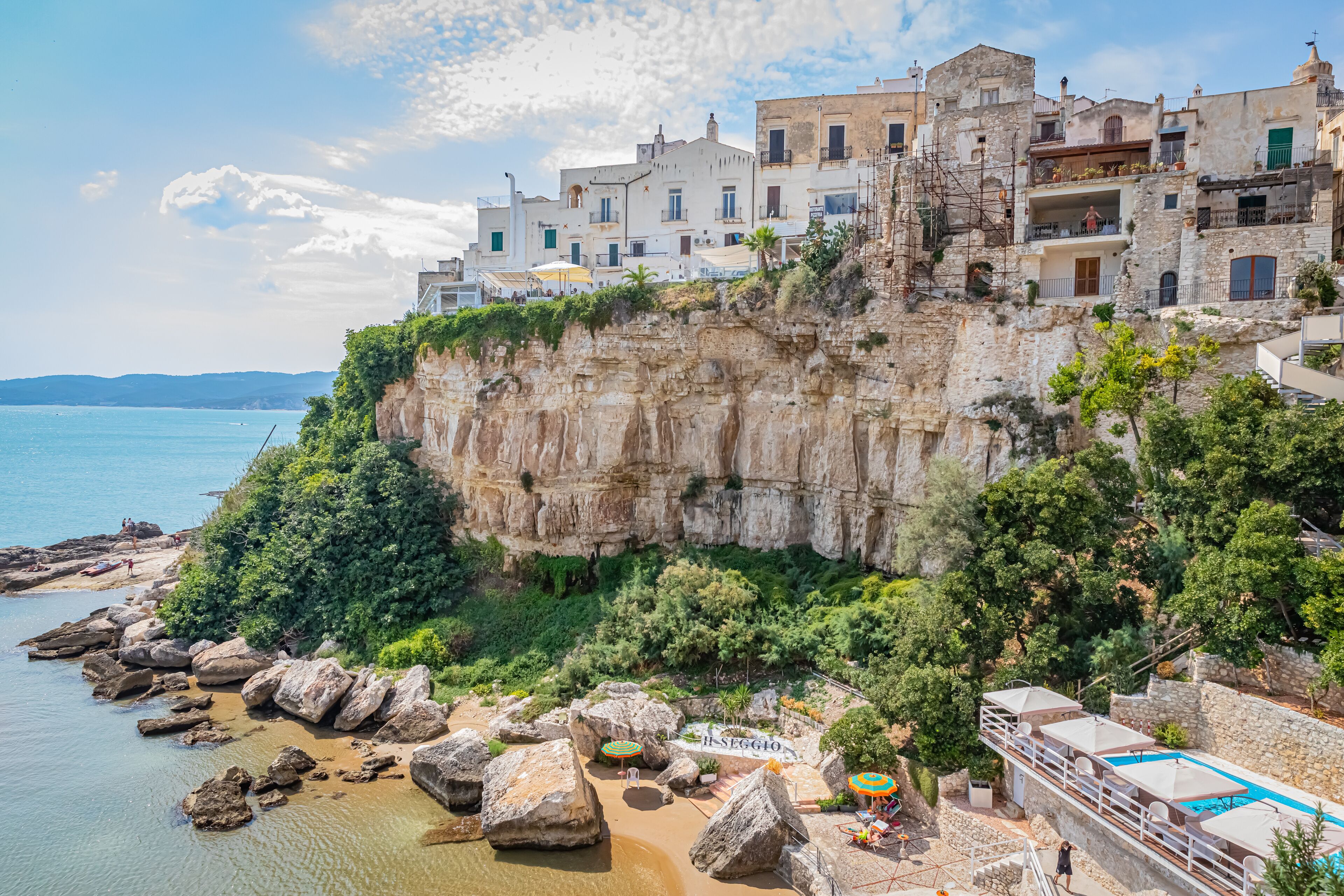 A coastal town with historic buildings perched on a cliff, a beach with large rocks, and a swimming pool.