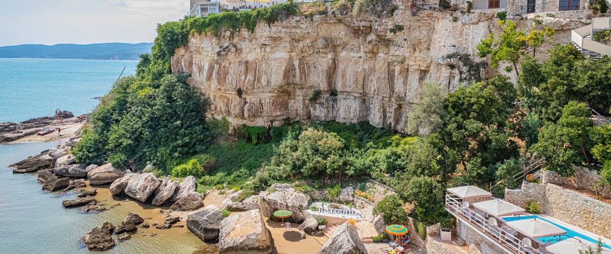 A coastal town with historic buildings perched on a cliff, a beach with large rocks, and a swimming pool.