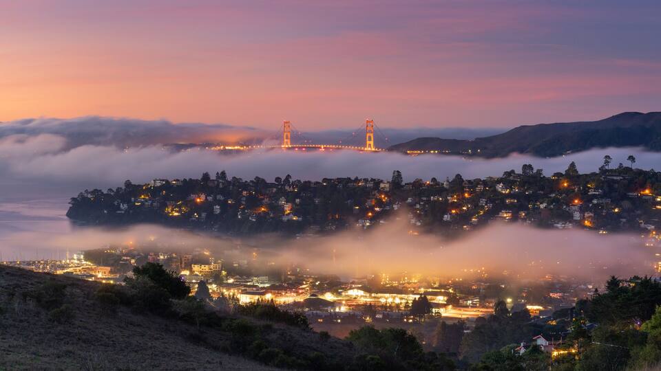 Un pueblo costero al atardecer con un puente al fondo y niebla sobre el agua.