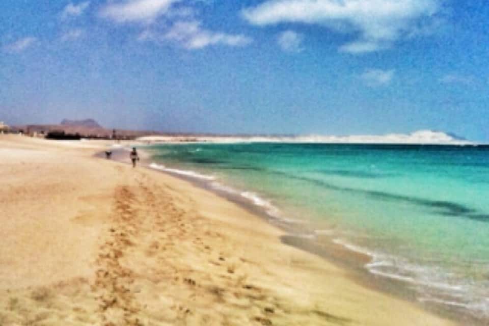 Praia arenosa com pegadas que levam até a água, céu claro e montanhas ao longe.