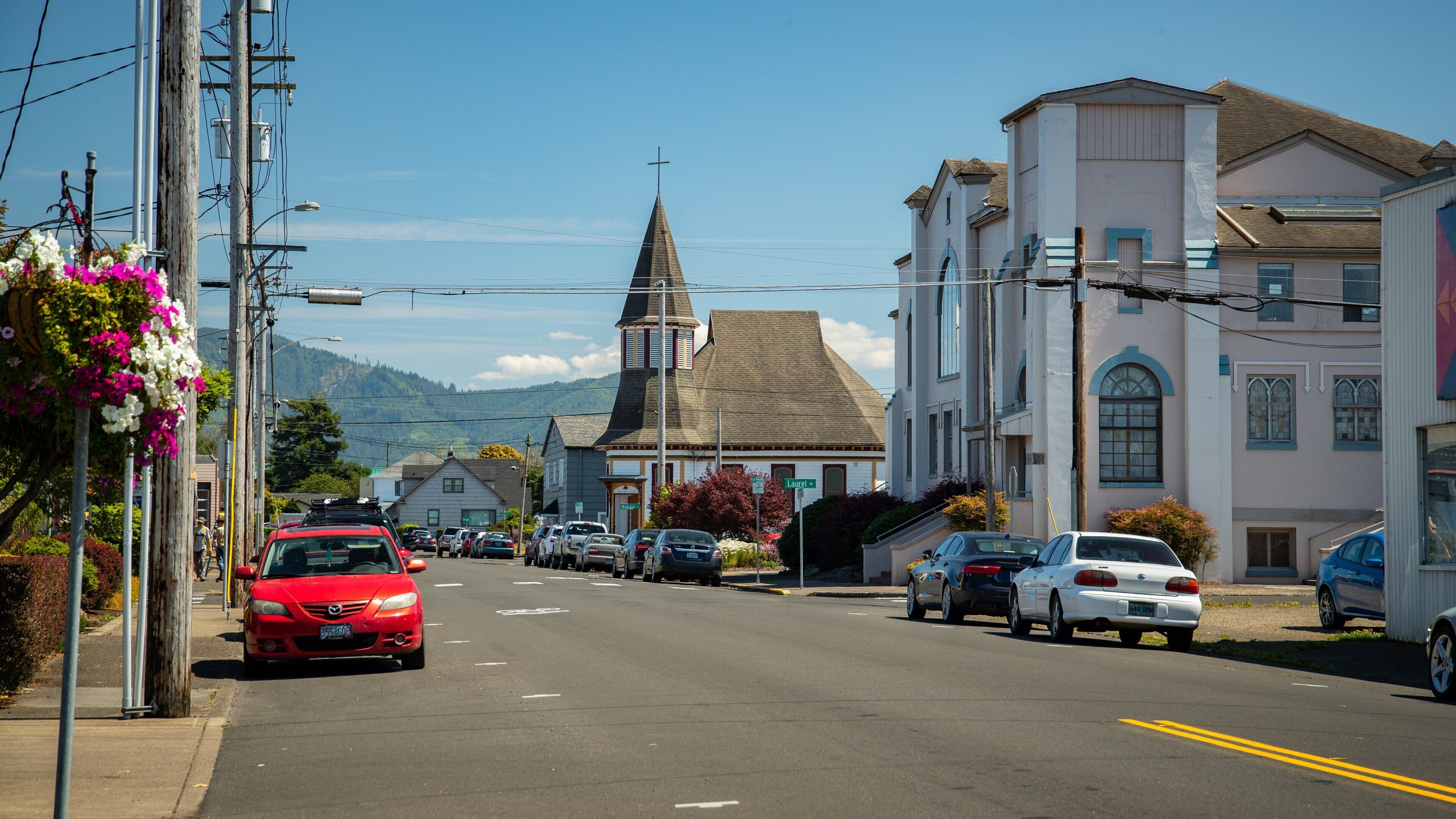 A street with parked cars, a church with a steeple, and buildings with bay windows.