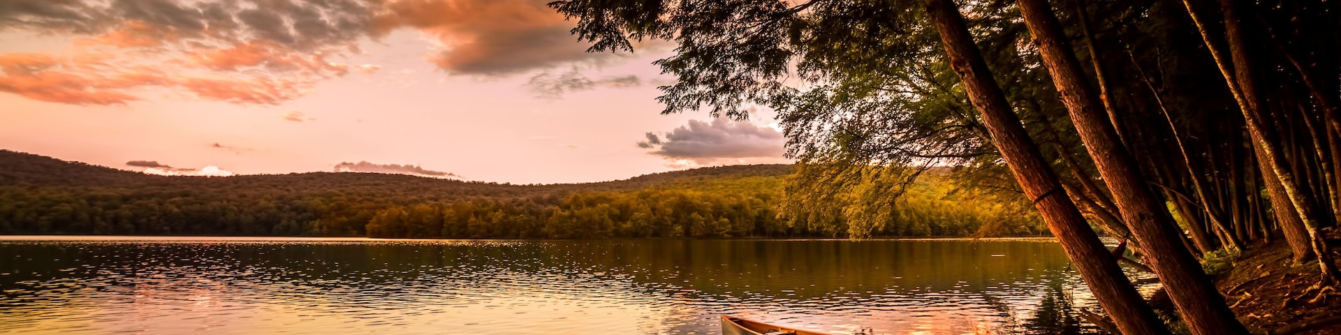 A calm lake at sunset with a canoe tied to a dock, surrounded by trees and hills.