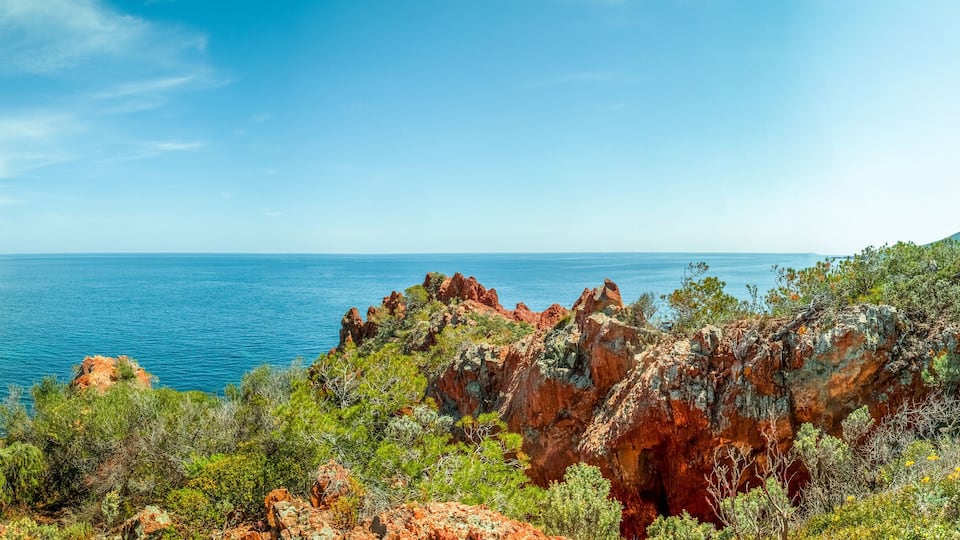 A coastal landscape with red cliffs, green vegetation, and a clear blue sea.