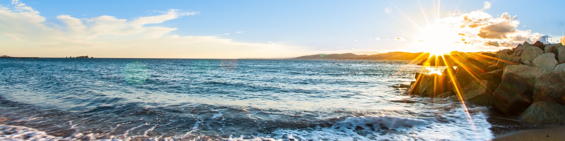 Una playa con olas, cielo despejado y el sol poniéndose en el horizonte.