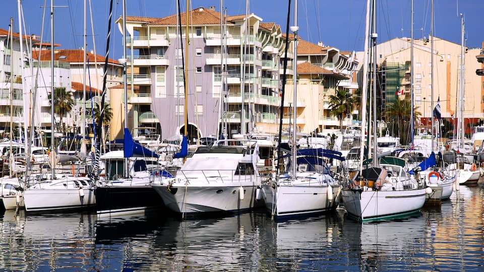 A marina with several sailboats docked, surrounded by multi-story buildings with balconies and a clear blue sky.