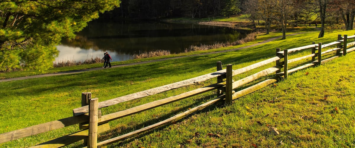 A lake, a wooden fence, a hilly landscape, and a person walking.