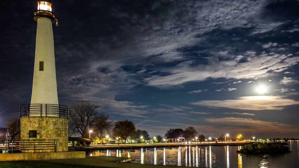A lighthouse by a calm body of water at night with a full moon in the sky.