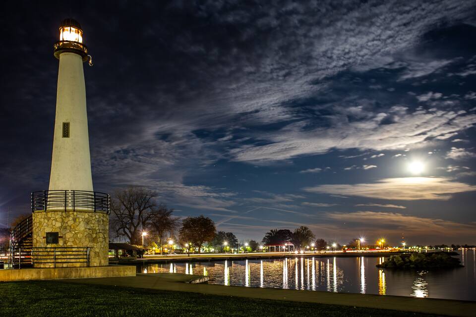 A lighthouse by a calm body of water at night with a full moon in the sky.