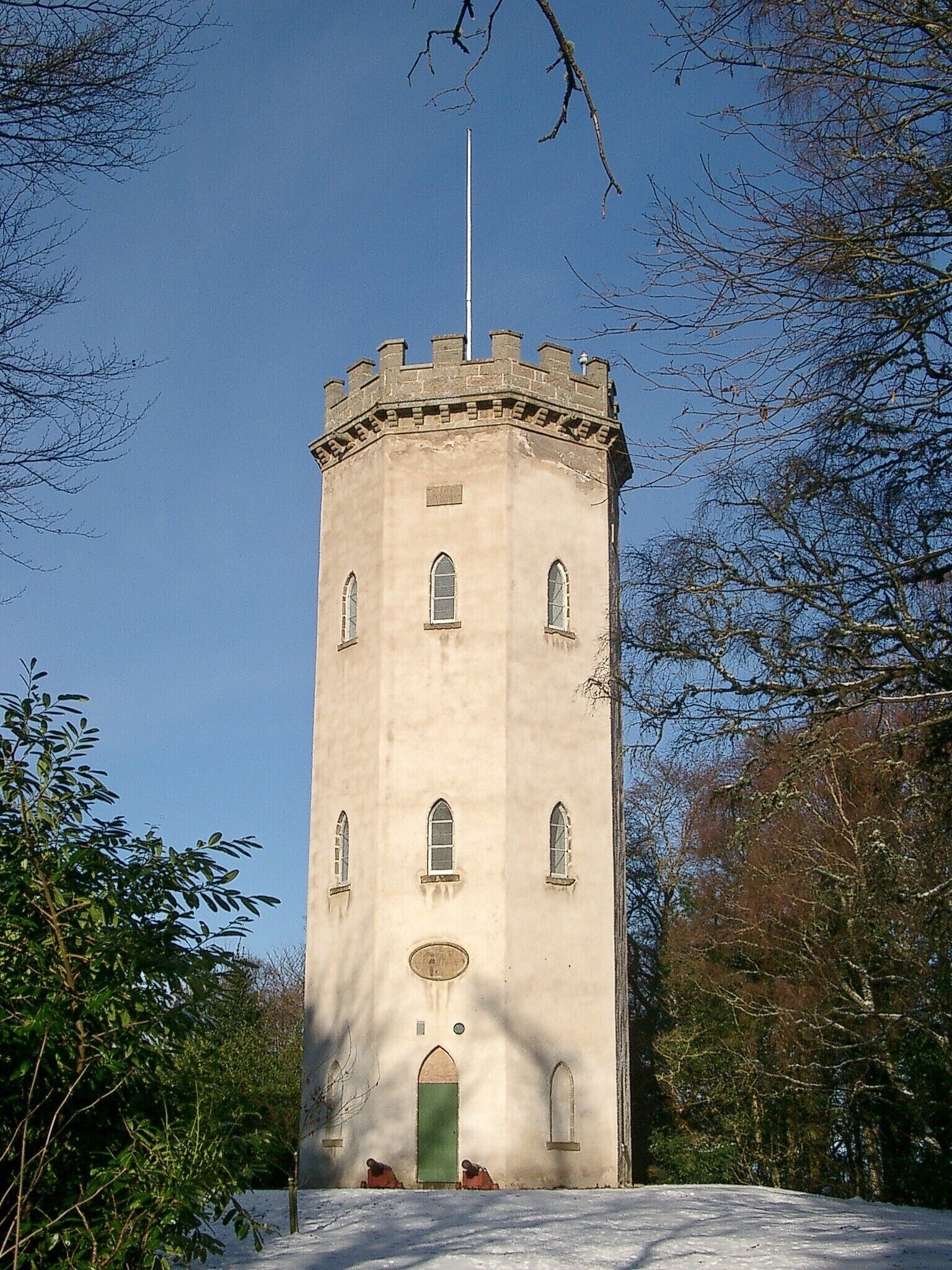 Ein hoher, beiger Turm mit Bogenfenstern und einer Fahnenstange, umgeben von Bäumen und Schnee.