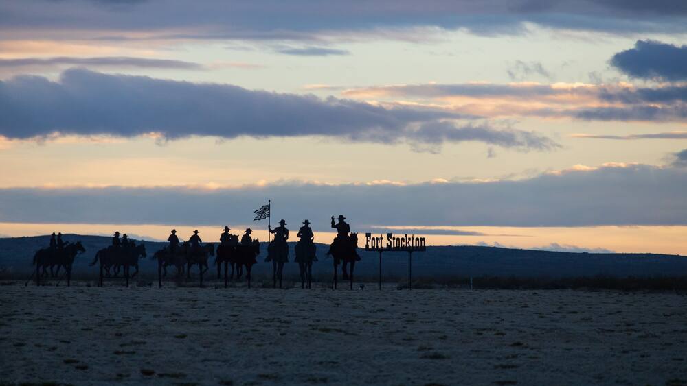 A group of people on horseback at dusk with a sign reading 'Fort Stockton' in the background.