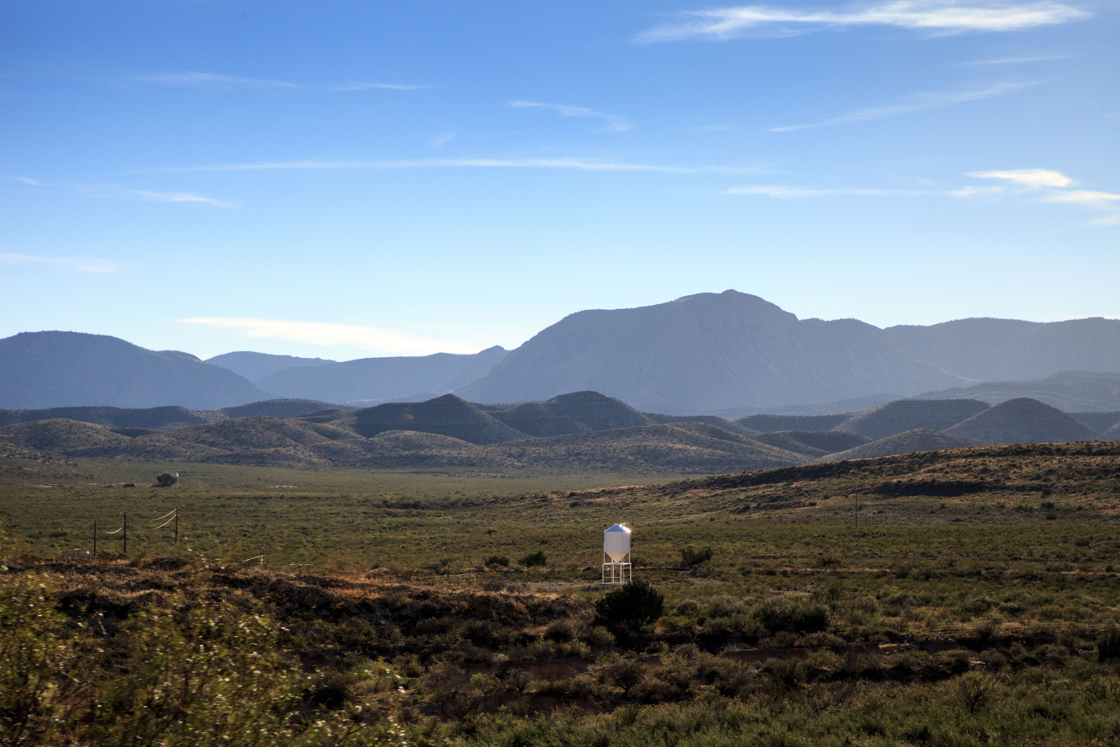 A white water tower in a grassy field with rolling hills and mountains in the background.
