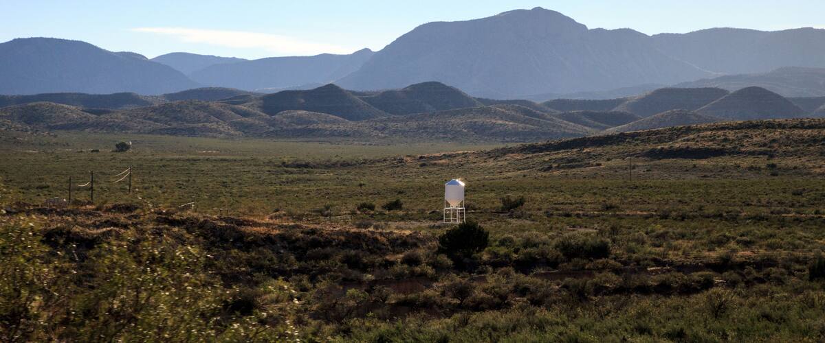 A white water tower in a grassy field with rolling hills and mountains in the background.