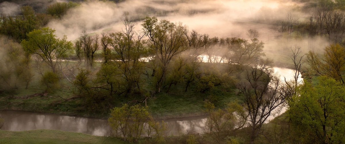 A misty river valley with trees and hills during sunrise.