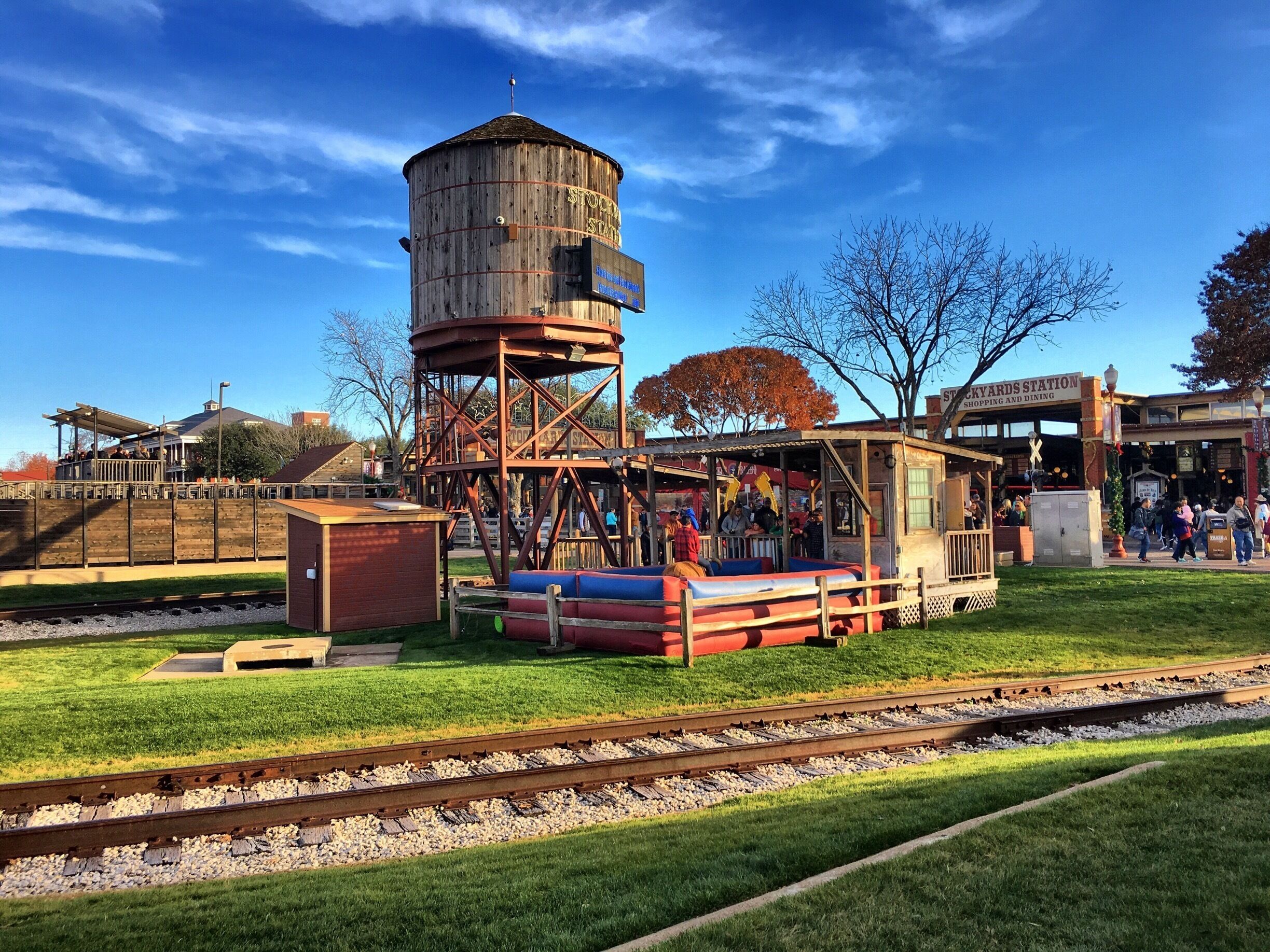 Una estación de tren antigua con una torre de agua de madera, mesas de picnic y un pequeño edificio con el rótulo 'Stockyards Station'.