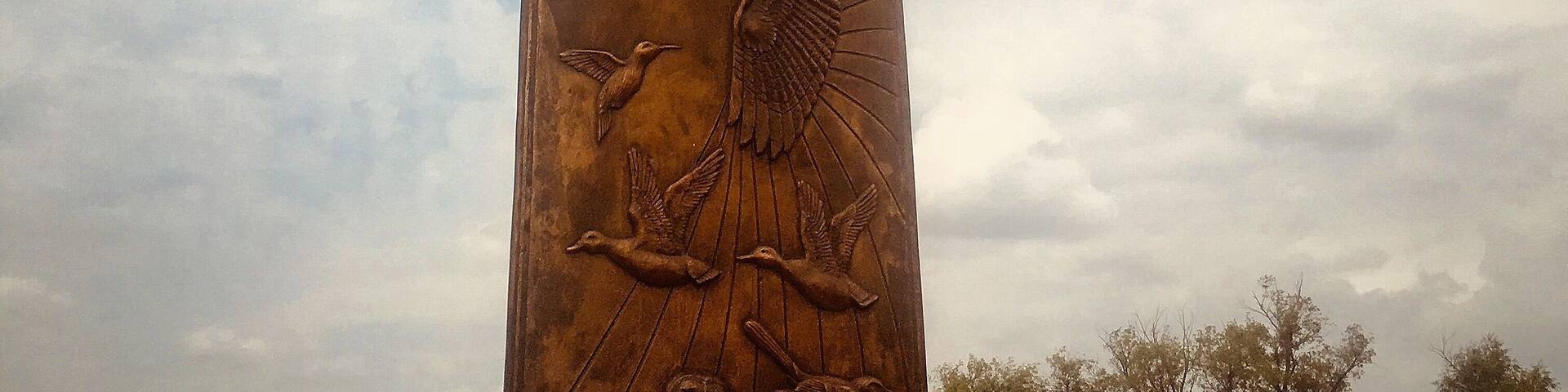 A bronze sculpture of a man with outstretched arms, a bird, and a fish, set against a cloudy sky.