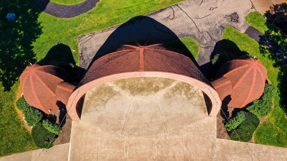 An aerial view of a large, arched structure with a red roof, surrounded by green lawns and a paved area.