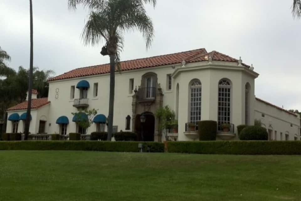 A large white building with a red-tiled roof, surrounded by a well-maintained lawn and palm trees.