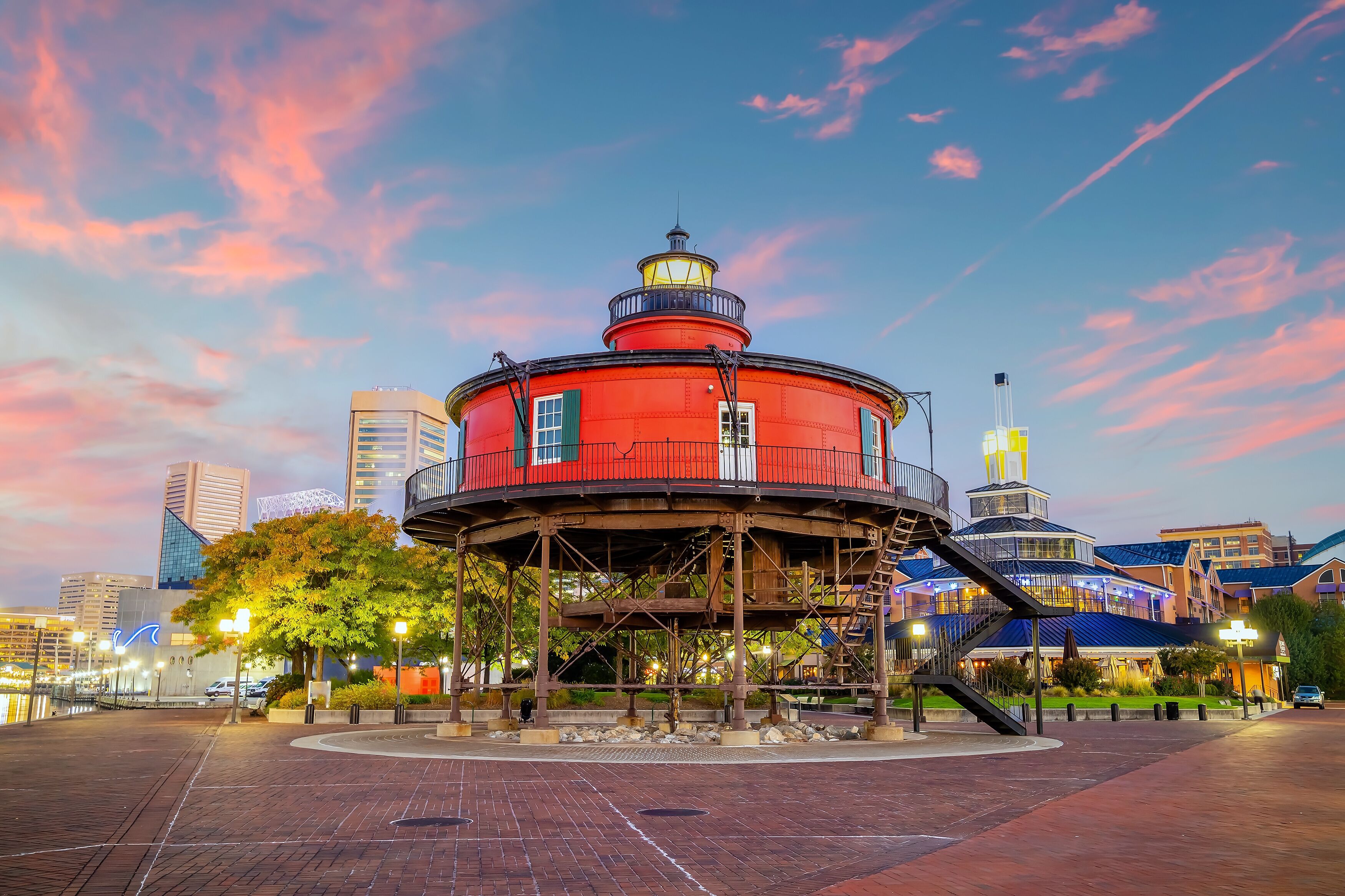 A red lighthouse with a balcony, surrounded by a brick plaza with trees and modern buildings in the background.
