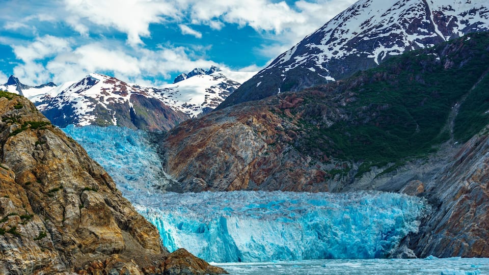 A glacier with a turquoise hue, surrounded by rugged mountains and snow-capped peaks under a partly cloudy sky.
