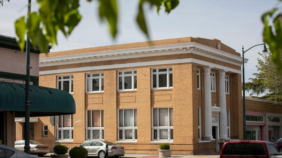A brick building with white trim and columns, surrounded by parked cars and a green awning.