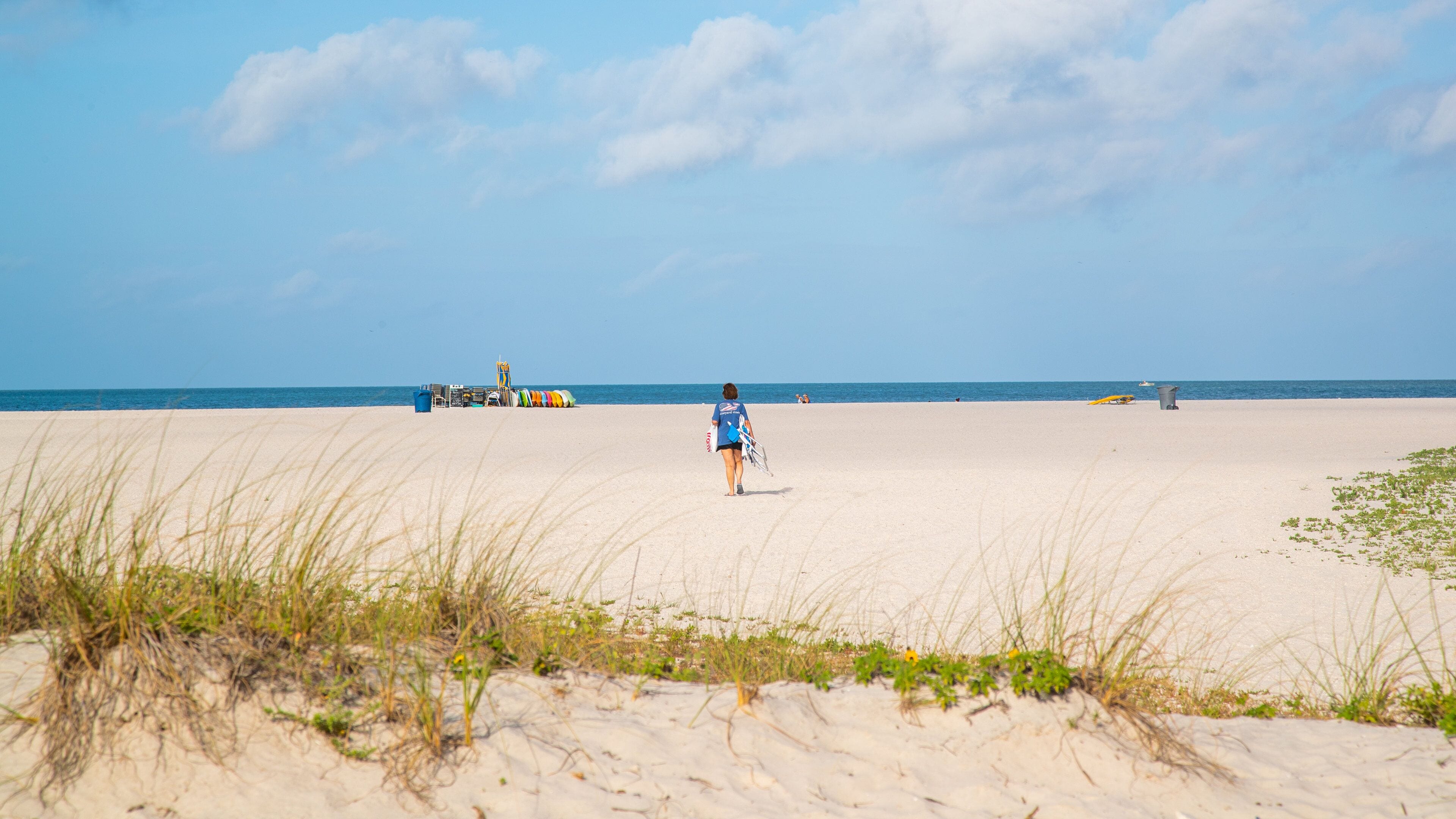 A sandy beach with a person walking towards the ocean, carrying a surfboard. There are beach umbrellas and a lifeguard tower in the background.