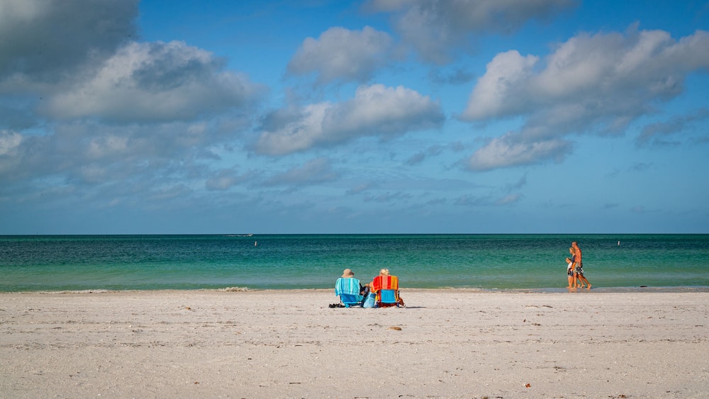 A beach with two people sitting on towels, a woman walking, and a boat in the distance.