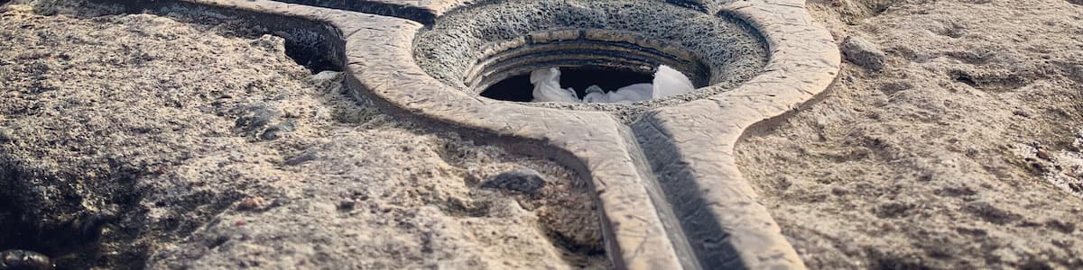 Un monument de pierre avec une structure en forme de croix au premier plan et un petit bâtiment surmonté d’un drapeau à l’arrière-plan.