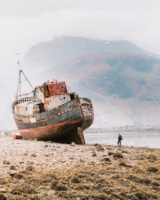 A rusted shipwreck on a rocky shore with a person standing nearby and a mountain in the background.