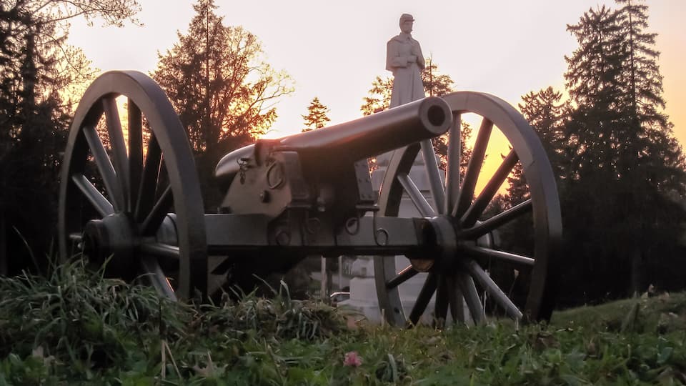 A cannon in a grassy area with trees and a statue in the background.