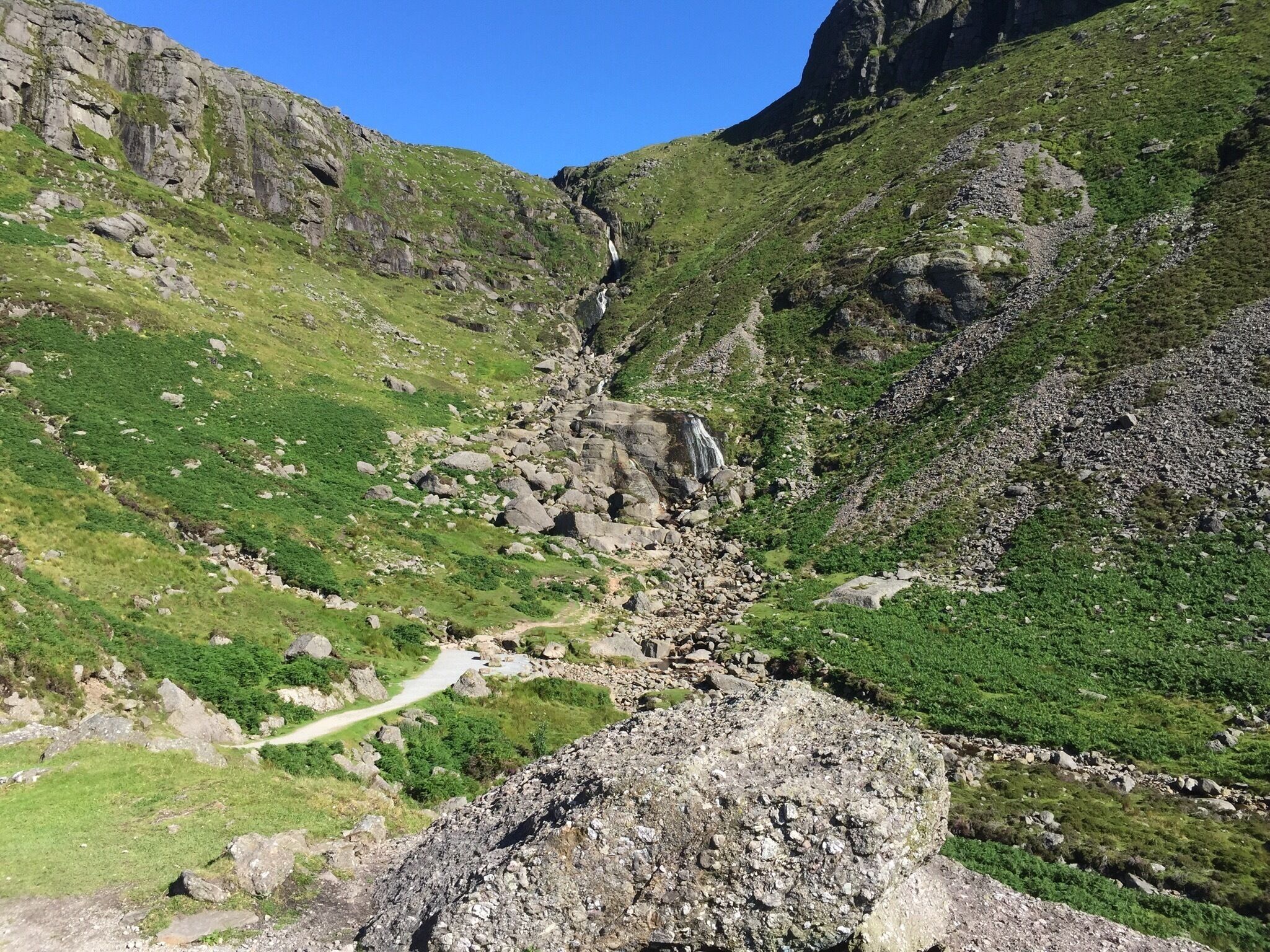 A mountainous landscape with a rocky path, green vegetation, and a waterfall.