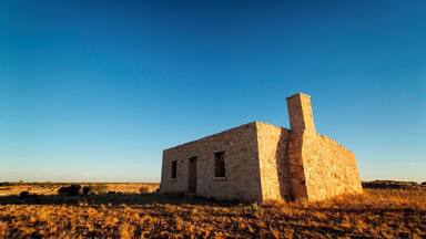 Ein altes Steinhaus mit Schornstein in einem Feld mit trockenem Gras unter blauem Himmel.