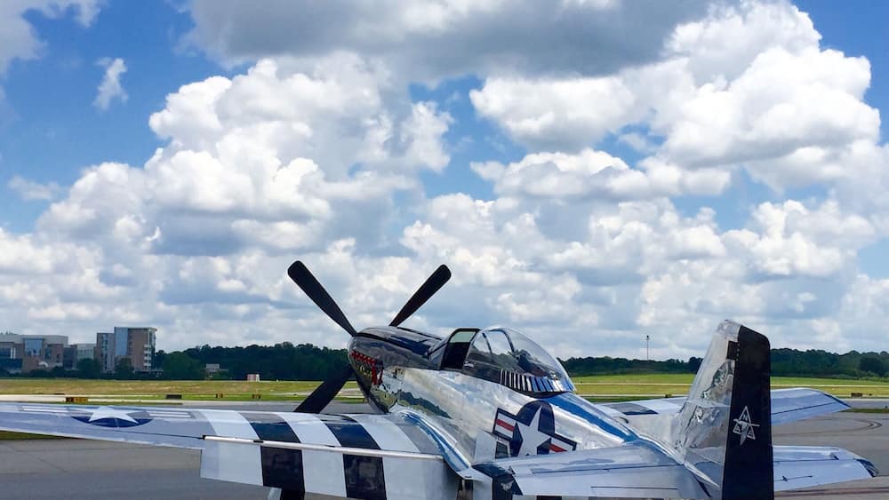 A vintage military aircraft with a propeller, a star insignia, and a checkered tail fin on a tarmac under a partly cloudy sky.