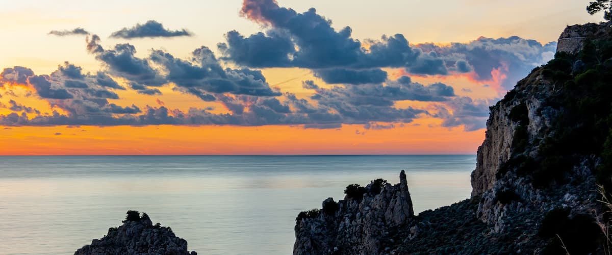 A rocky coastline at sunset with a calm sea and a sky with scattered clouds.