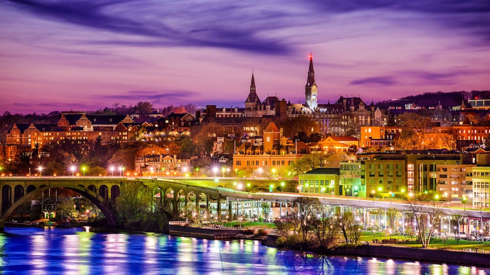 A cityscape at dusk with a prominent bridge over a river, illuminated buildings, and a tall tower with a spire.