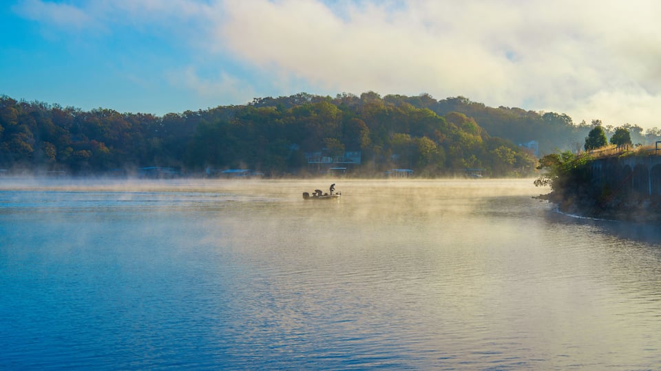 A calm lake with a boat carrying two people, surrounded by mist and a forested hillside.