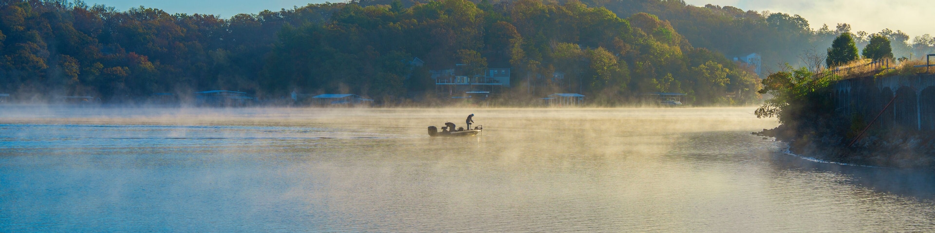 A calm lake with a boat carrying two people, surrounded by mist and a forested hillside.