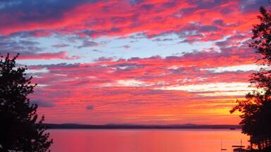A sunset over a calm lake with vibrant red and pink clouds, surrounded by trees and boats.