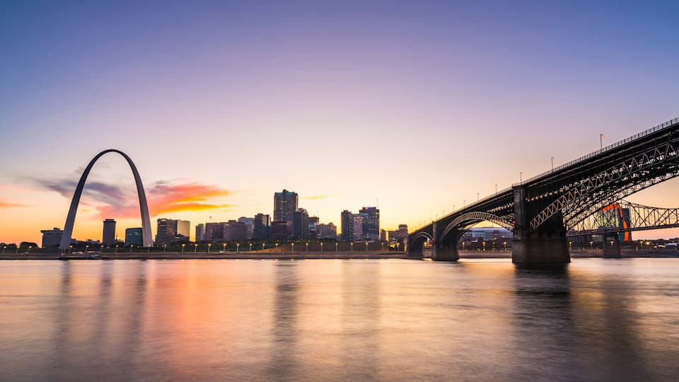 A city skyline at dusk with a prominent arch bridge and a river reflecting the sky.