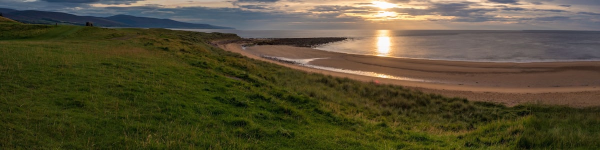 Un paysage côtier avec une colline herbeuse, une côte rocheuse et un coucher de soleil sur l’océan.